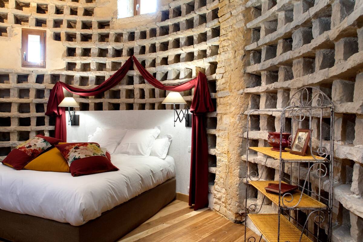 Un dormitorio con pared de piedra, cama con ropa de cama blanca, almohadas rojas y amarillas, y estante metálico con adornos.