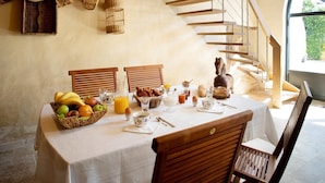 A breakfast table set with fruits, pastries, and beverages, overlooking a stone staircase and a large window.