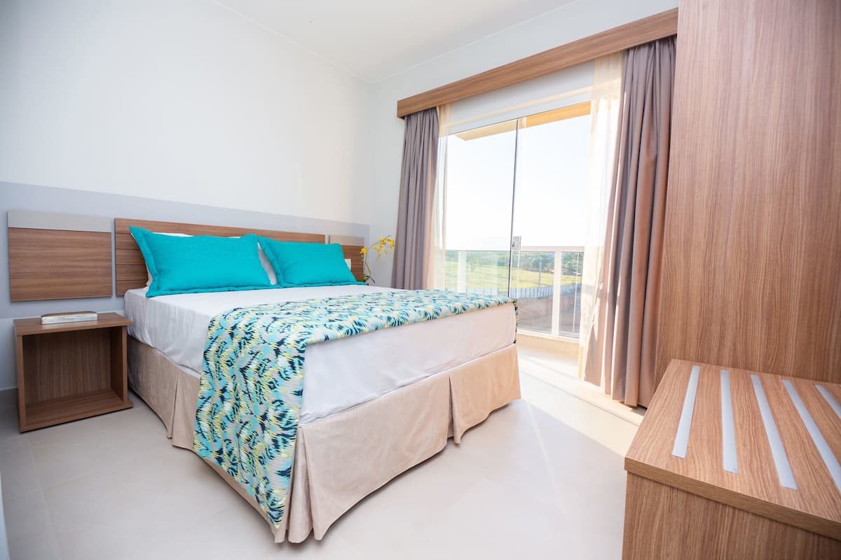 A modern hotel room with a large bed, a wooden nightstand, a small table, and a balcony with curtains.