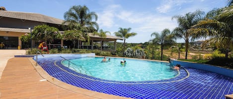 Une piscine aux carreaux bleus, entourée de palmiers et bordée d’un bâtiment dont le toit est recouvert de tuiles.