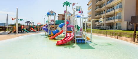 A water park with multiple slides and play structures, surrounded by a fence and buildings in the background.