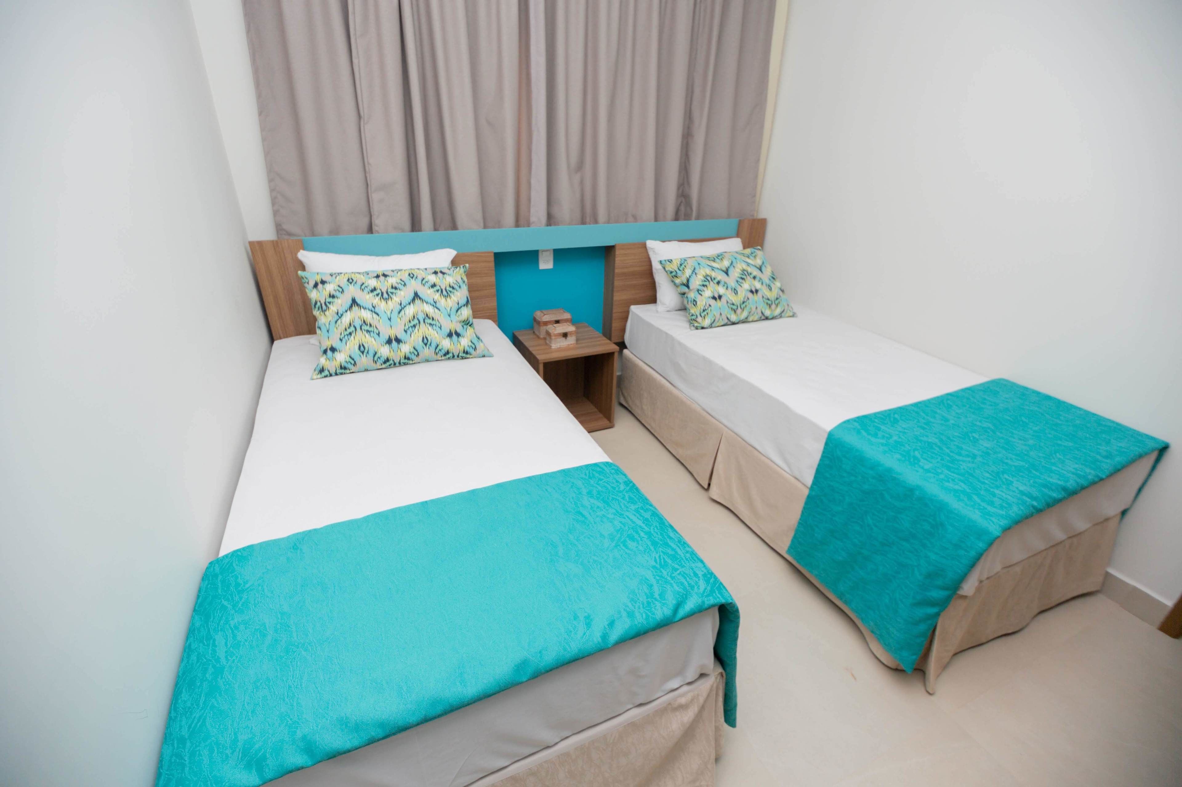 A hotel room with two single beds, each with a teal bedspread and matching pillows. There is a wooden nightstand between the beds, and a small wooden block on it. The room has a simple, clean design with a light-colored floor and a curtain on the window.