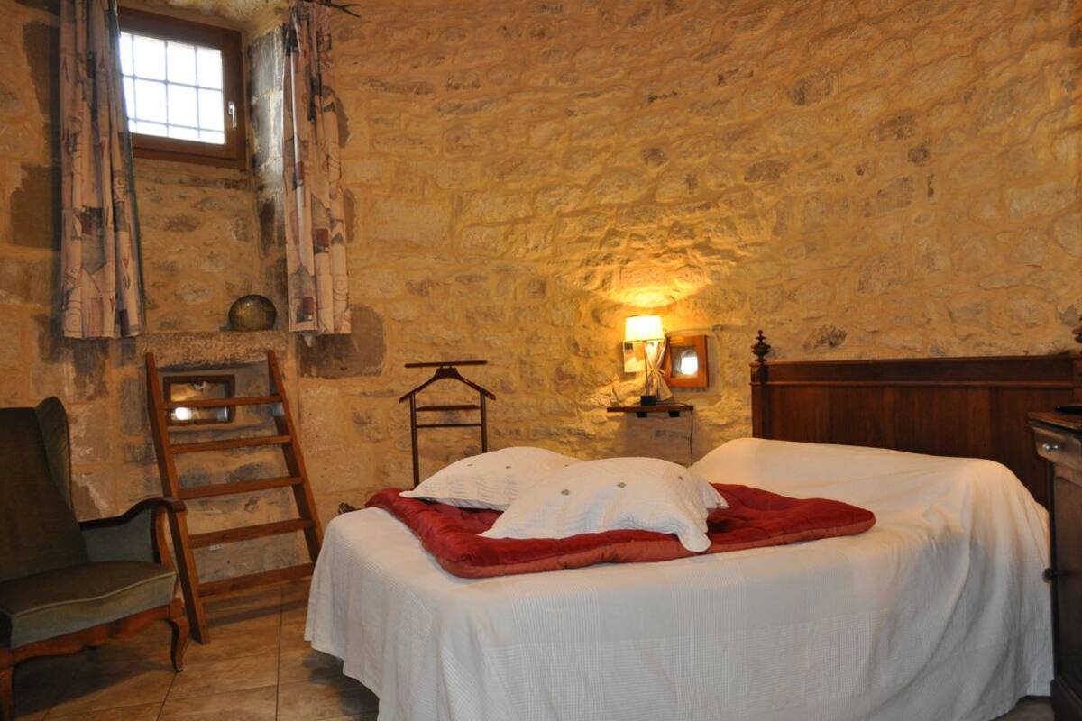 Une pièce avec un mur en pierre, un lit avec une literie blanche et une couverture rouge, une chaise en bois, une échelle et une petite table avec une lampe.