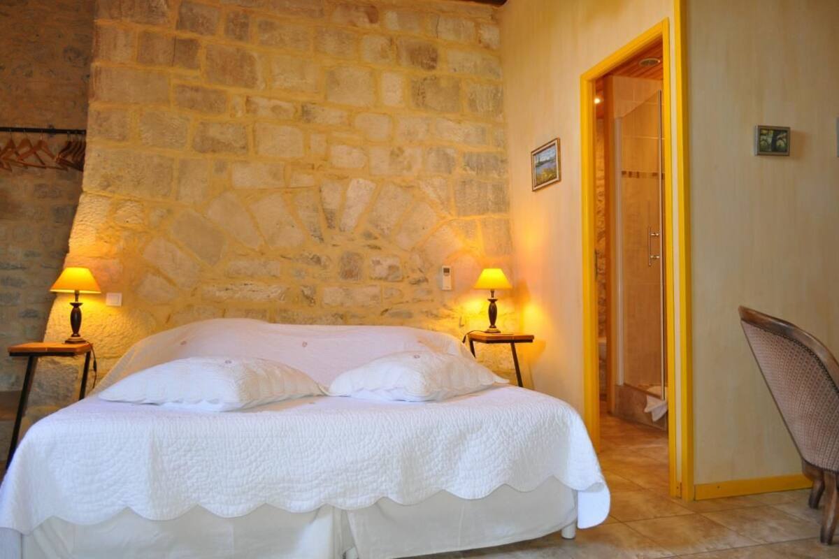 Une chambre avec un mur en pierre, un lit avec une literie blanche, deux lampes de chevet et une chaise.