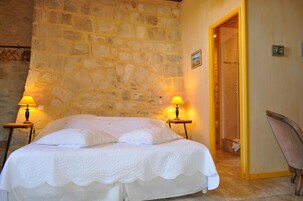 Un dormitorio con pared de piedra, una cama con ropa de cama blanca, dos lámparas de noche y una silla.