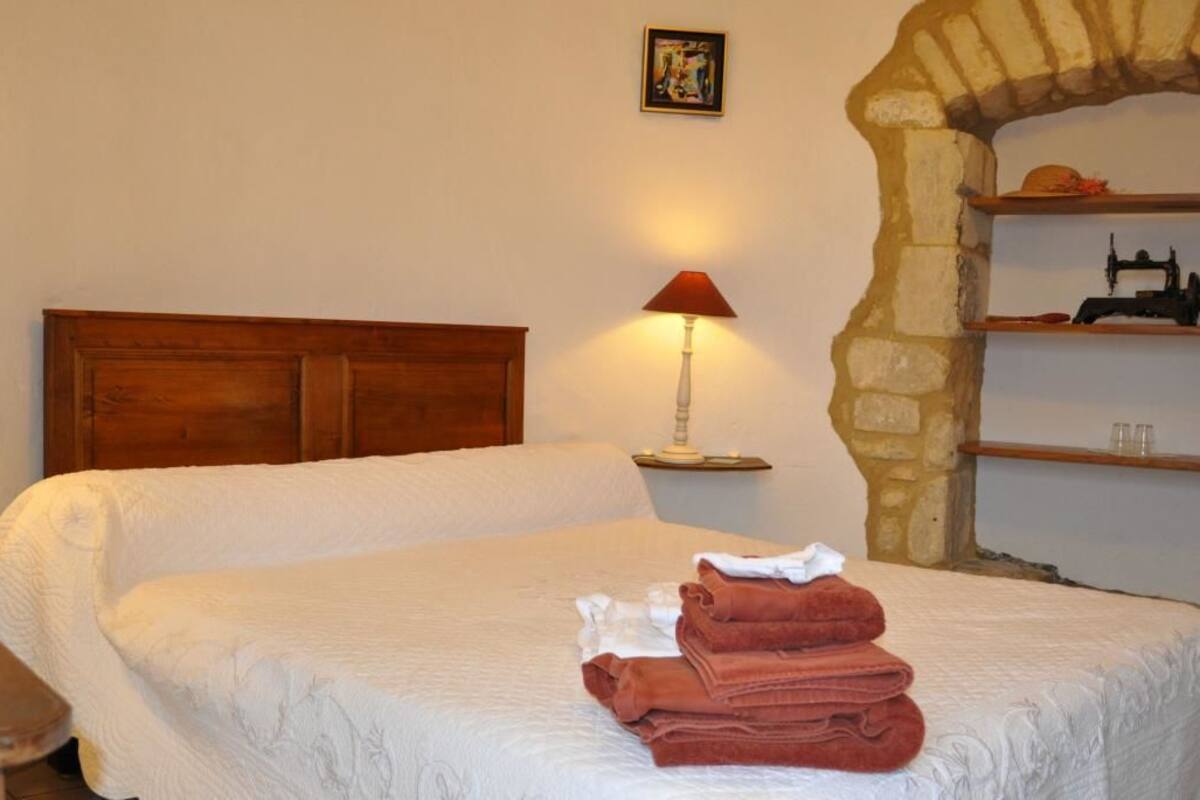 Un lit bien fait, avec une couette blanche et des serviettes rouges, deux lampes de chevet, une tête de lit en bois, une étagère en pierre sur laquelle se trouve une machine à coudre, et un petit tableau accroché au mur.