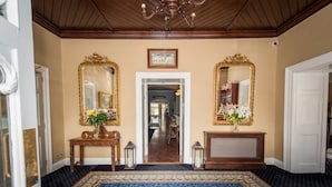 A grand hallway with ornate gold-framed mirrors, a wooden console table with a vase of flowers, and a patterned carpet leading to a dining area.
