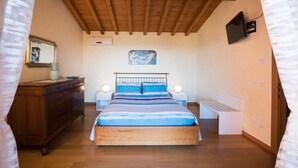 Un dormitorio con techo de madera, una cama con ropa de cama azul, un aparador de madera, lámparas de noche y un televisor montado en la pared.