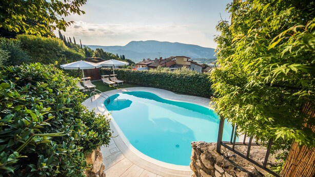 Una piscina rodeada de muros de piedra y exuberante vegetación, con vistas a montañas y casas a la distancia.