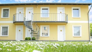 A yellow building with multiple windows and doors, a central spiral staircase, and a small garden with white flowers. - Appartementhaus Gutenberg - Für Paare, Freundinnen & Freunde, Alleinreisende - Parkplatz & WLAN - 2 Min zum See, 6 Min zum Strand (Binz)