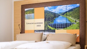 Una habitación de hotel con una cama grande, un cabecero que ofrece vistas panorámicas a un lago y montañas, y el texto "LINACHT ALSPERRE 1925".
