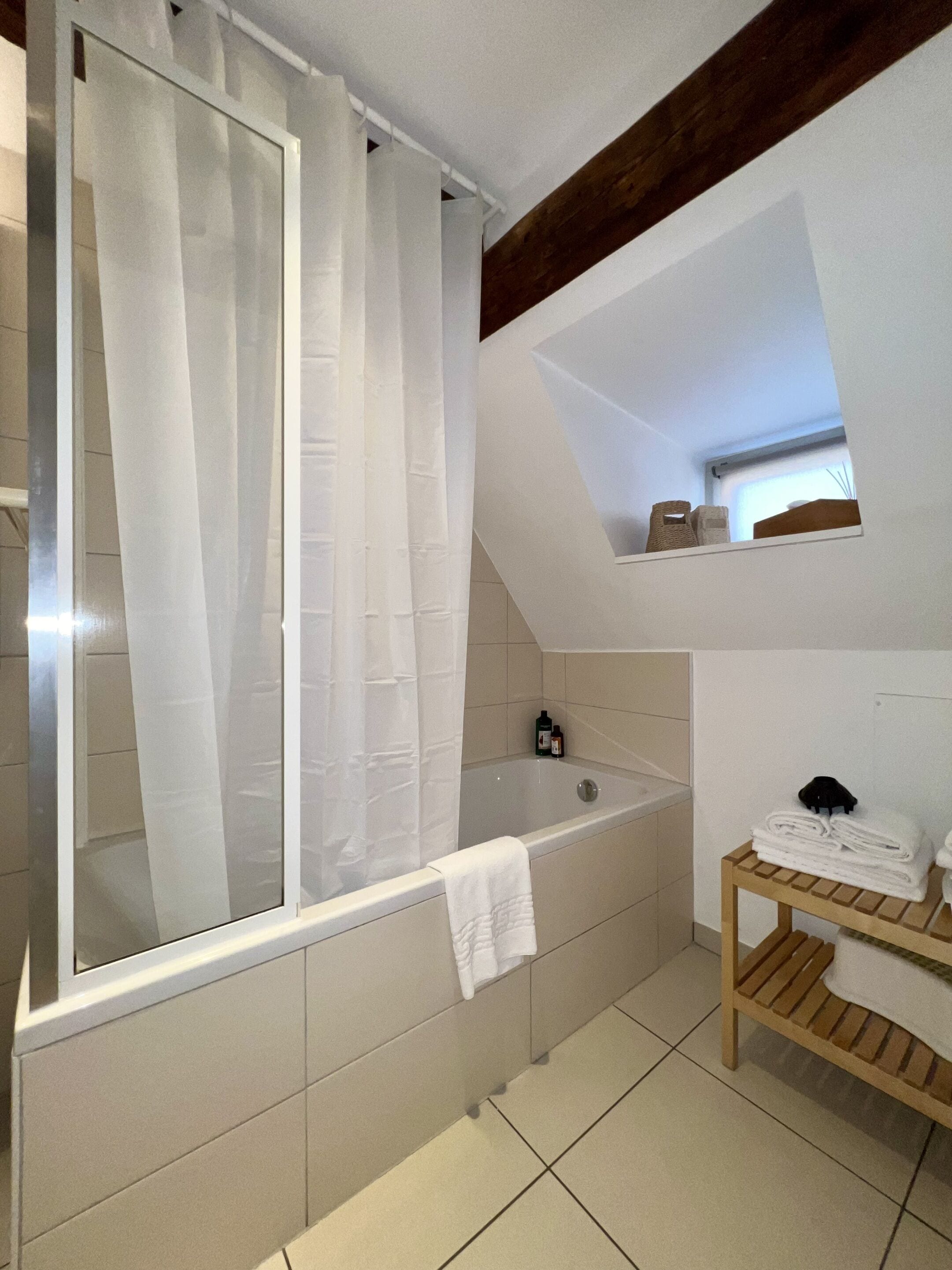 Une salle de bain avec une baignoire, un rideau de douche, une étagère en bois avec des serviettes et une petite lucarne.