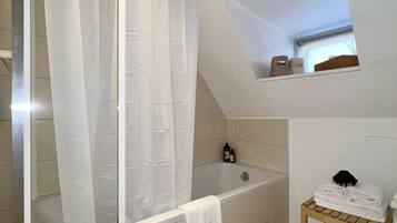 Une salle de bain avec une baignoire, un rideau de douche, une étagère en bois avec des serviettes et une petite lucarne.