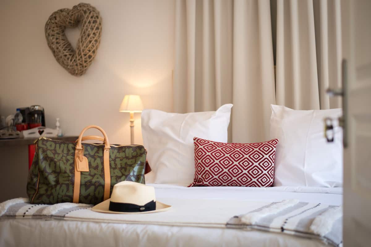 Een net opgemaakt bed met witte lakens, een gestreept kussen, een hoed en een Louis Vuitton tas.