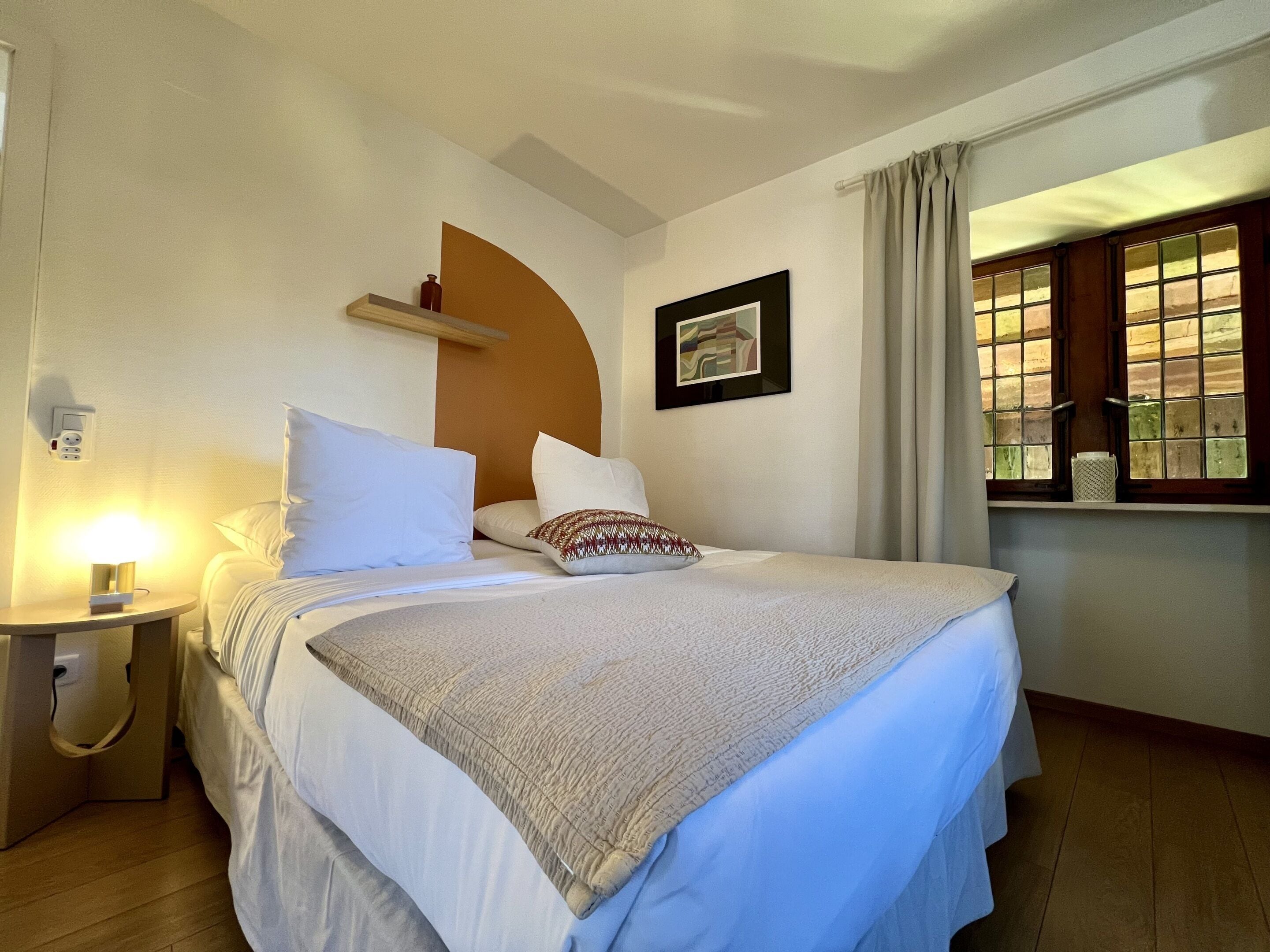 Une chambre d’hôtel avec un lit, une table de chevet, une lampe et une fenêtre avec des rideaux.