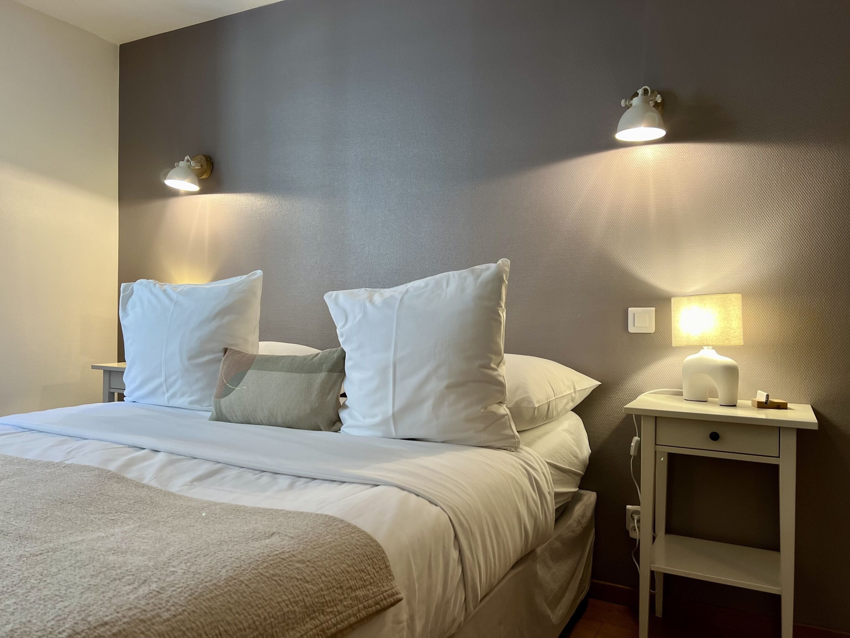 Ein ordentlich bezogenes Bett mit weißer Bettwäsche und einer beige farbenen Tagesdecke, ein Nachttisch mit Lampe sowie eine Wandleuchte.
