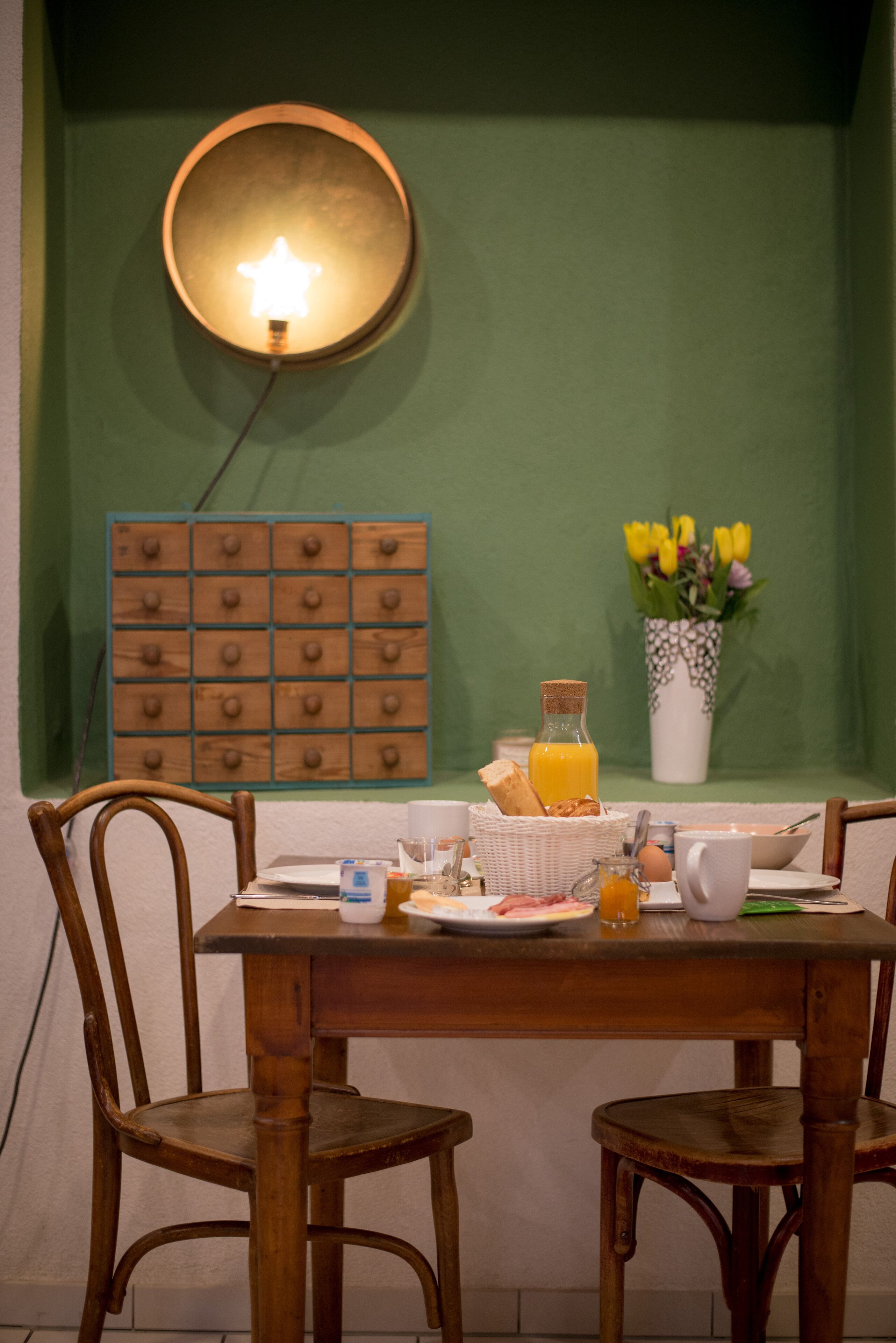 A dining table with breakfast items, a wall-mounted light, a flower vase, and a wooden panel with knobs.
