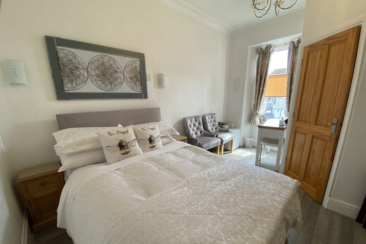 Quarto com cama grande, duas almofadas decorativas, cabeceira, mesa de cabeceira, cadeira, pequena mesa com abajur, janela com cortinas e porta de madeira.
