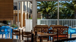 A poolside area with wooden chairs and tables, a white railing, and a view of palm trees and a building in the background. - Hôtel Leukos (Leucate)