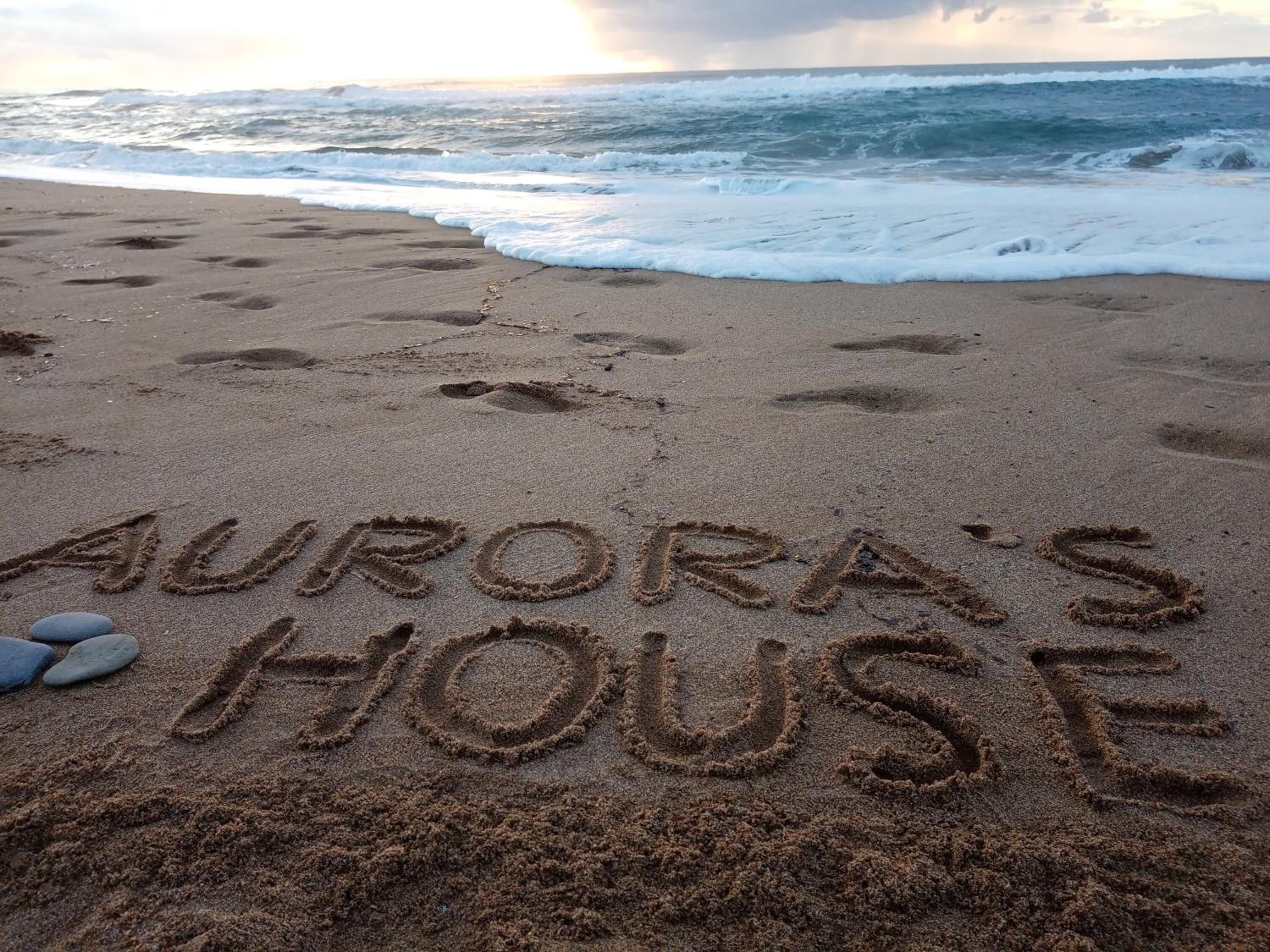 Una playa con 'AURORA'S HOUSE' escrito en la arena, huellas y algunas piedras.