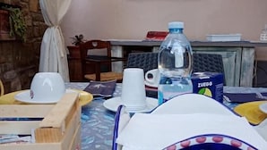 Una mesa dispuesta con una botella de agua, vasos y un bolso, con una ventana y un muro de ladrillos al fondo.