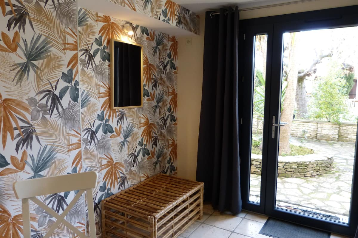 A room with a tropical leaf-patterned wall, a small table, a bench, and a view of an outdoor stone patio through a glass door.