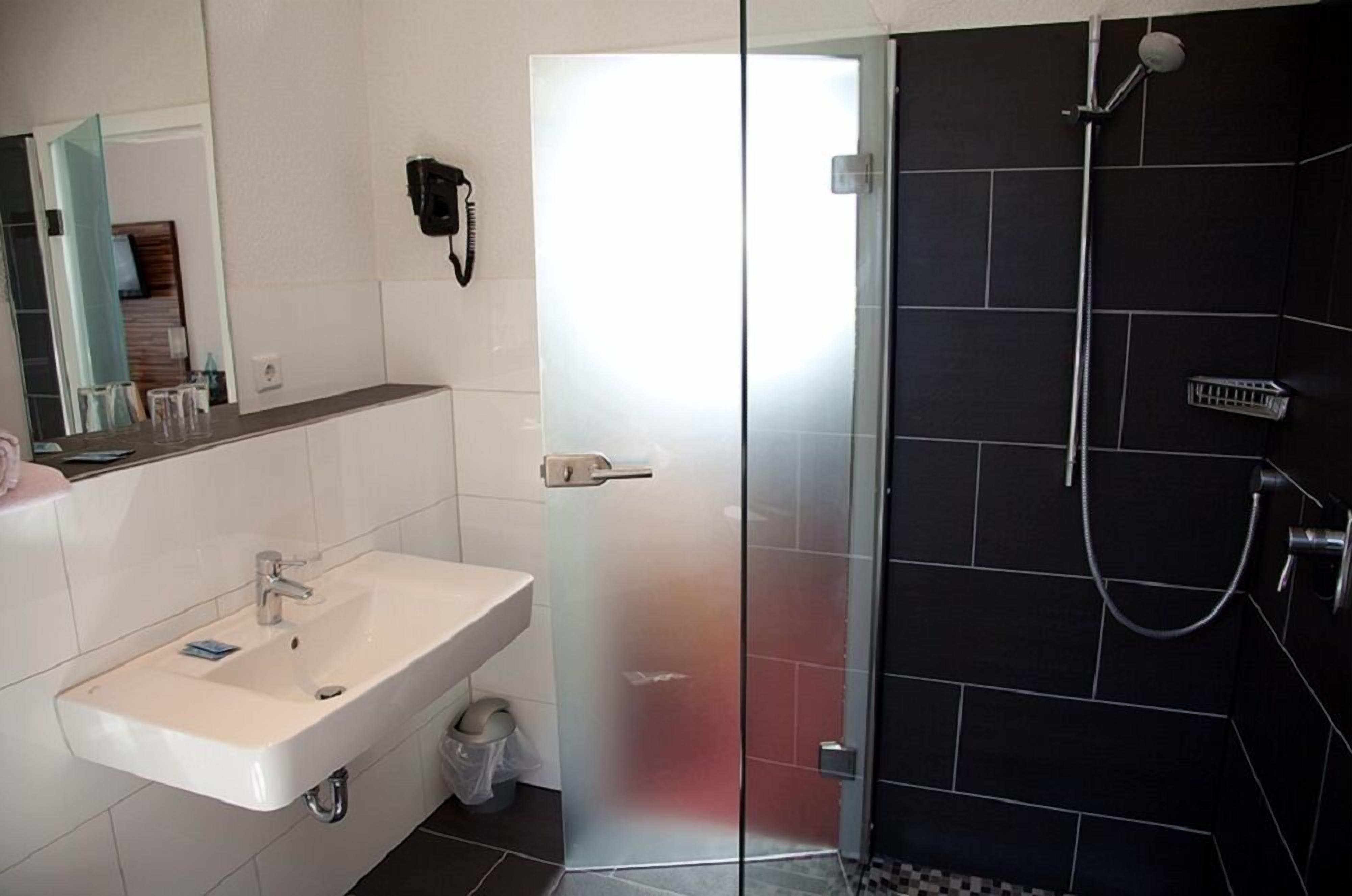 Ein modernes Badezimmer mit einem weißen Waschbecken, einer Duschkabine aus Glas und einem dunklen, gefliesten Duschbereich.