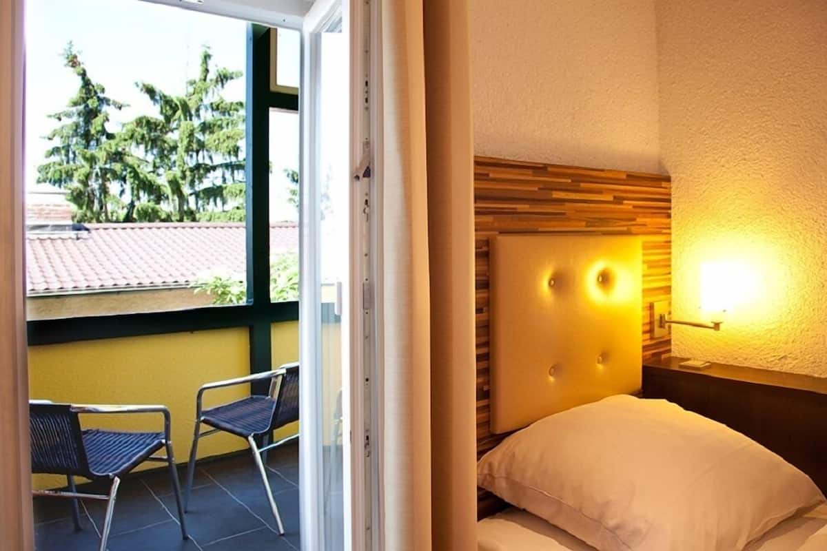 A hotel room with a bed, a headboard with lights, a window with a view of trees, and a balcony with chairs.