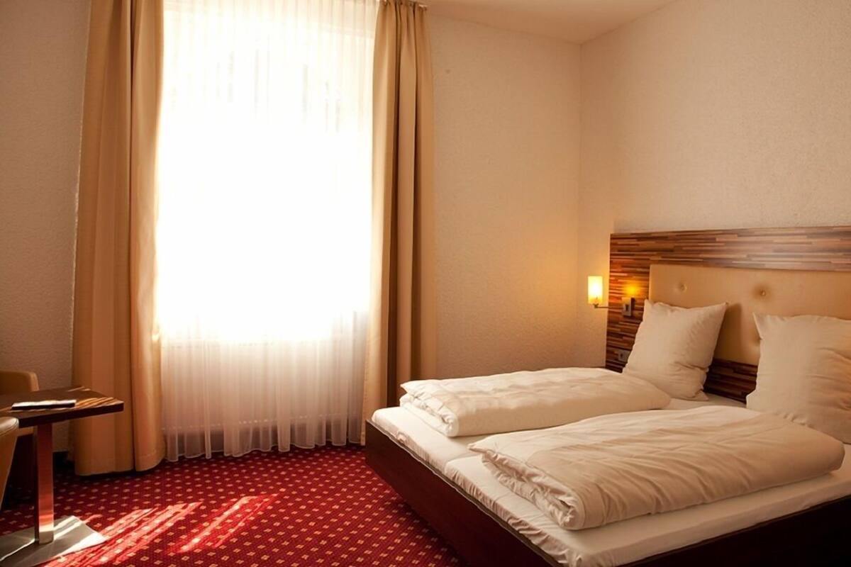 A hotel room with two beds, a desk, and a window with sheer curtains.