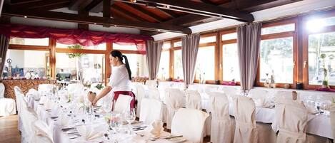 Un salón de banquetes con largas mesas dispuestas para un evento formal, una persona arreglando la vajilla, y amplias ventanas con cortinas.