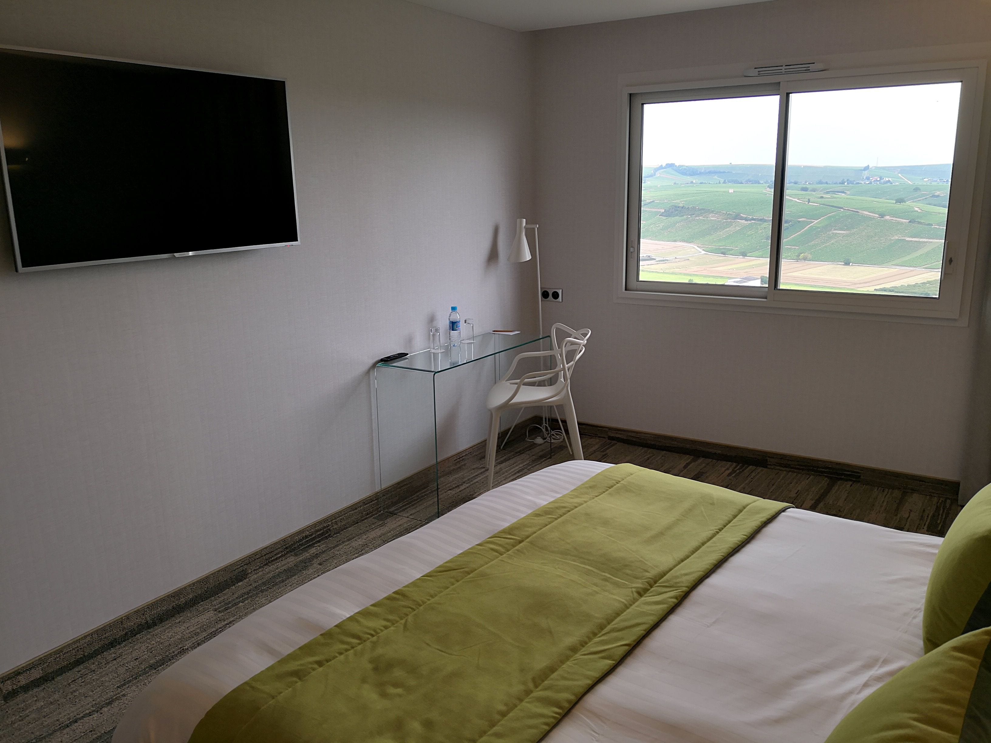 A hotel room with a bed, a TV mounted on the wall, a window with a view of fields, and a glass table with a chair.