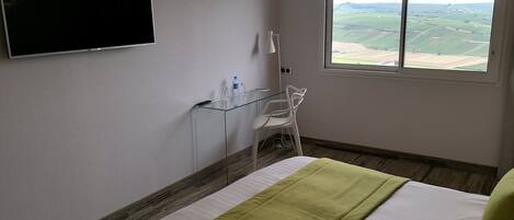 A hotel room with a bed, a TV mounted on the wall, a window with a view of fields, and a glass table with a chair.