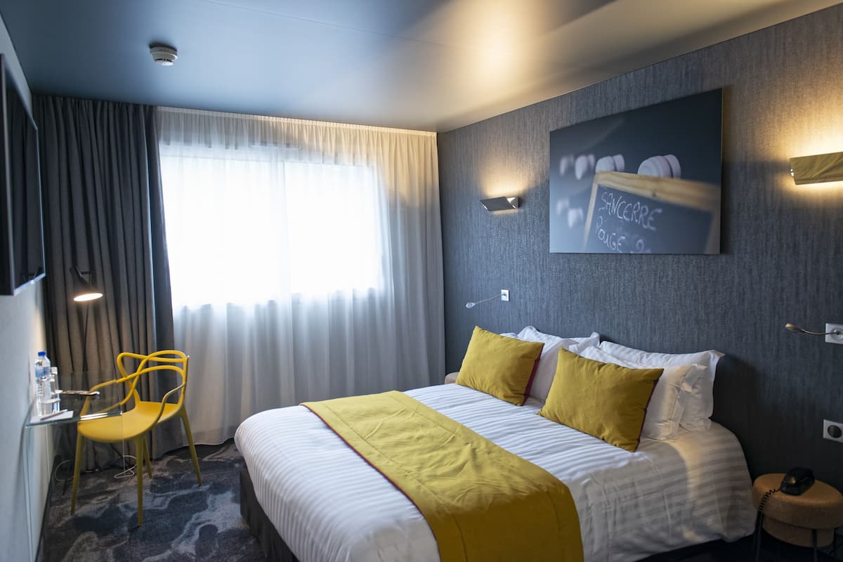 Habitación de hotel con cama, escritorio y silla, ventana con cortinas y una obra de arte en la pared.