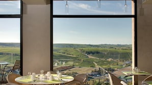 A dining area with a table set for two, large windows offering a view of a vineyard and rolling hills. - Le Panoramic Hôtel (Sancerre)