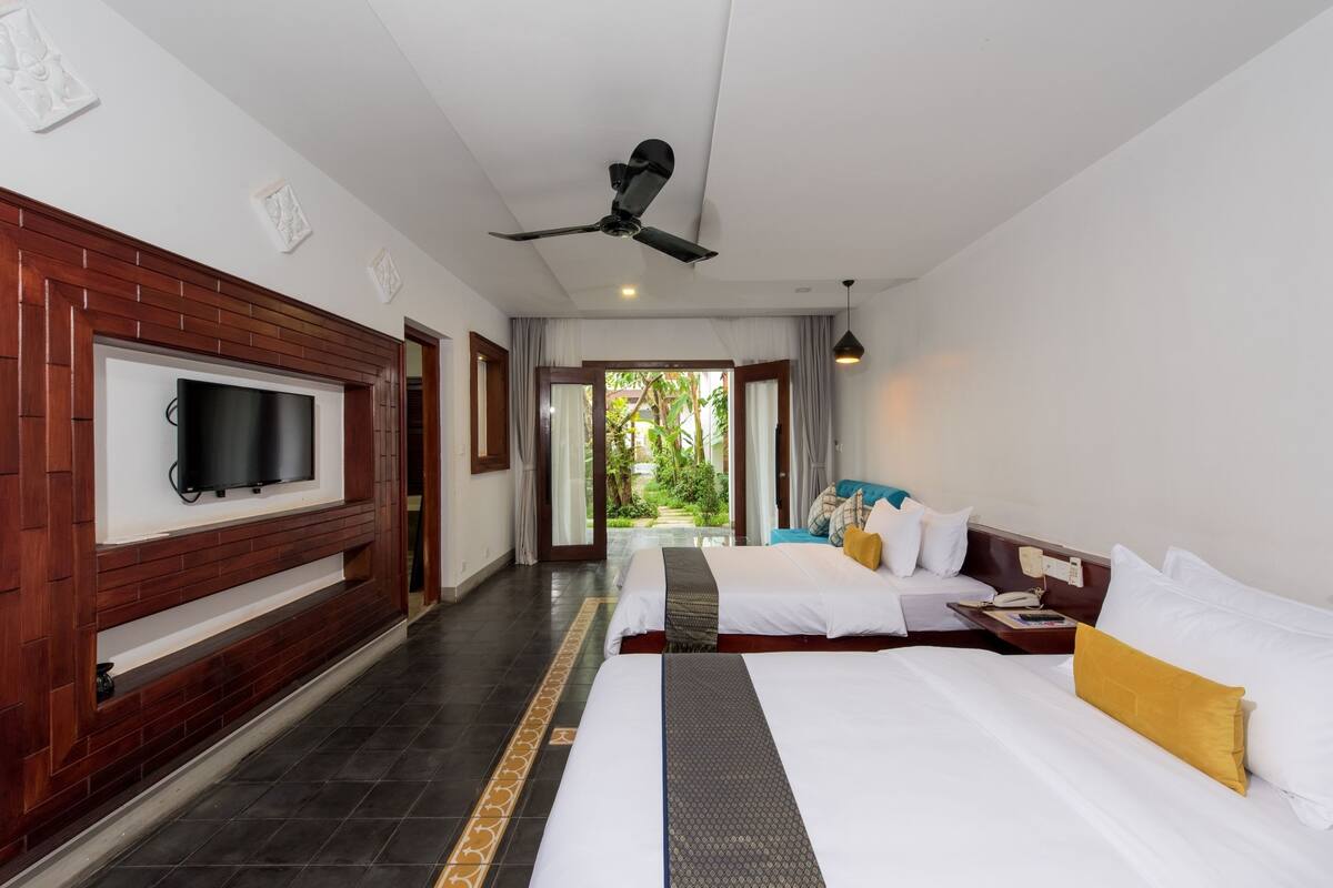 A hotel room with a TV mounted on a wooden panel, two beds with white bedding and yellow pillows, a ceiling fan, and a view of greenery through the door.