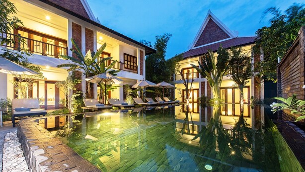 A poolside area with lounge chairs and umbrellas, surrounded by a two-story building with a tiled roof and a stone wall. - Charming Angkor Resort & Spa (Siem Reap)