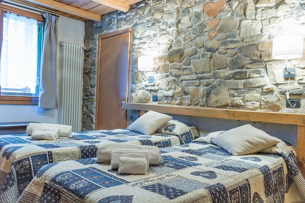 A bedroom with a stone wall, a wooden headboard, a window with curtains, and a bed with a patterned quilt and pillows.