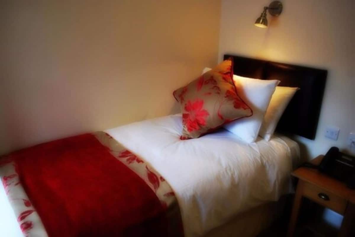 A neatly made bed with a white and red bedspread, a red throw blanket, and a floral pillow. There is a bedside table with a lamp and a small drawer.