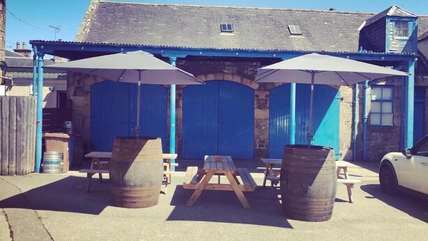 A rustic outdoor seating area with wooden tables and benches, barrels, and umbrellas. - The Aberlour Hotel (Aberlour)