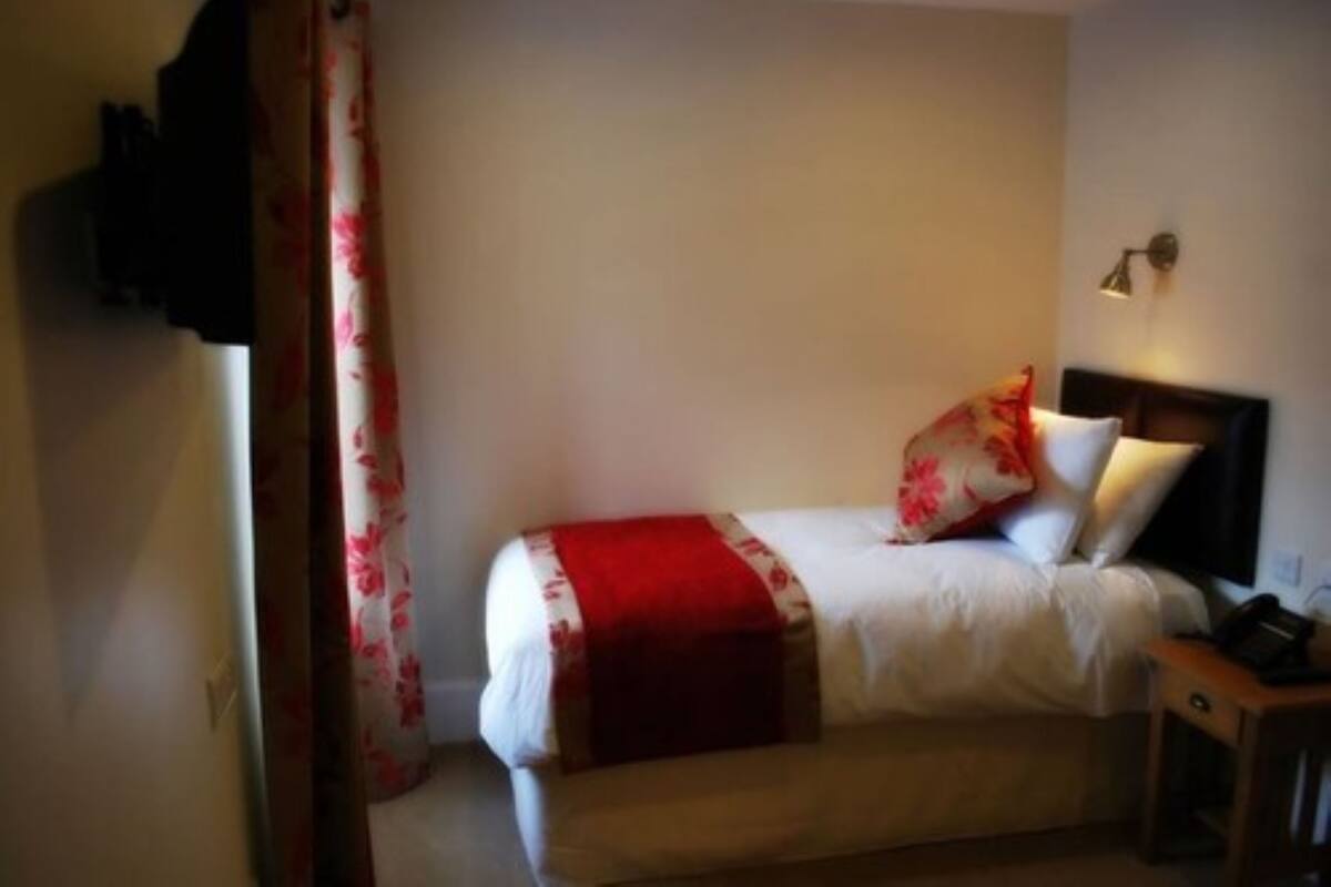 A single bed with a red and white blanket, a wooden nightstand, a lamp, and a curtain with a floral pattern.