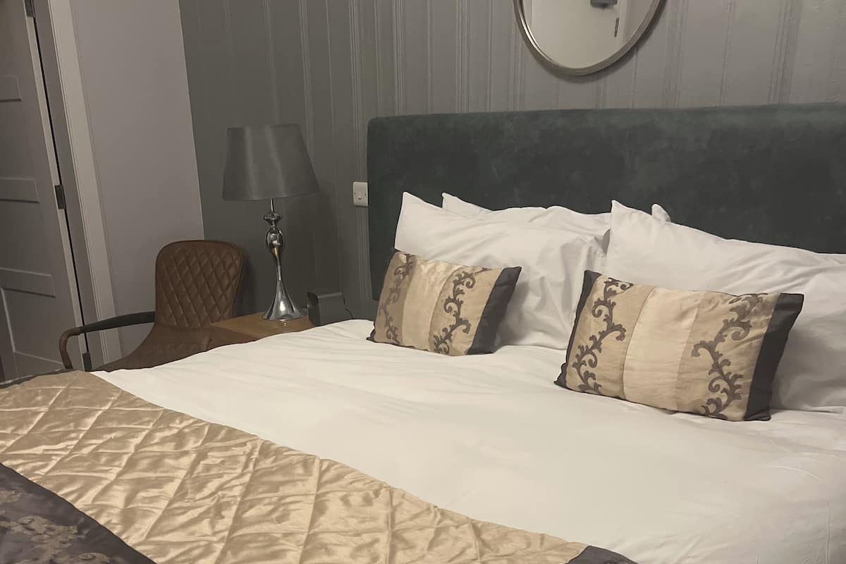 A neatly made bed with a patterned bedspread, a round mirror on the wall, and a chair with a lamp beside the bed.