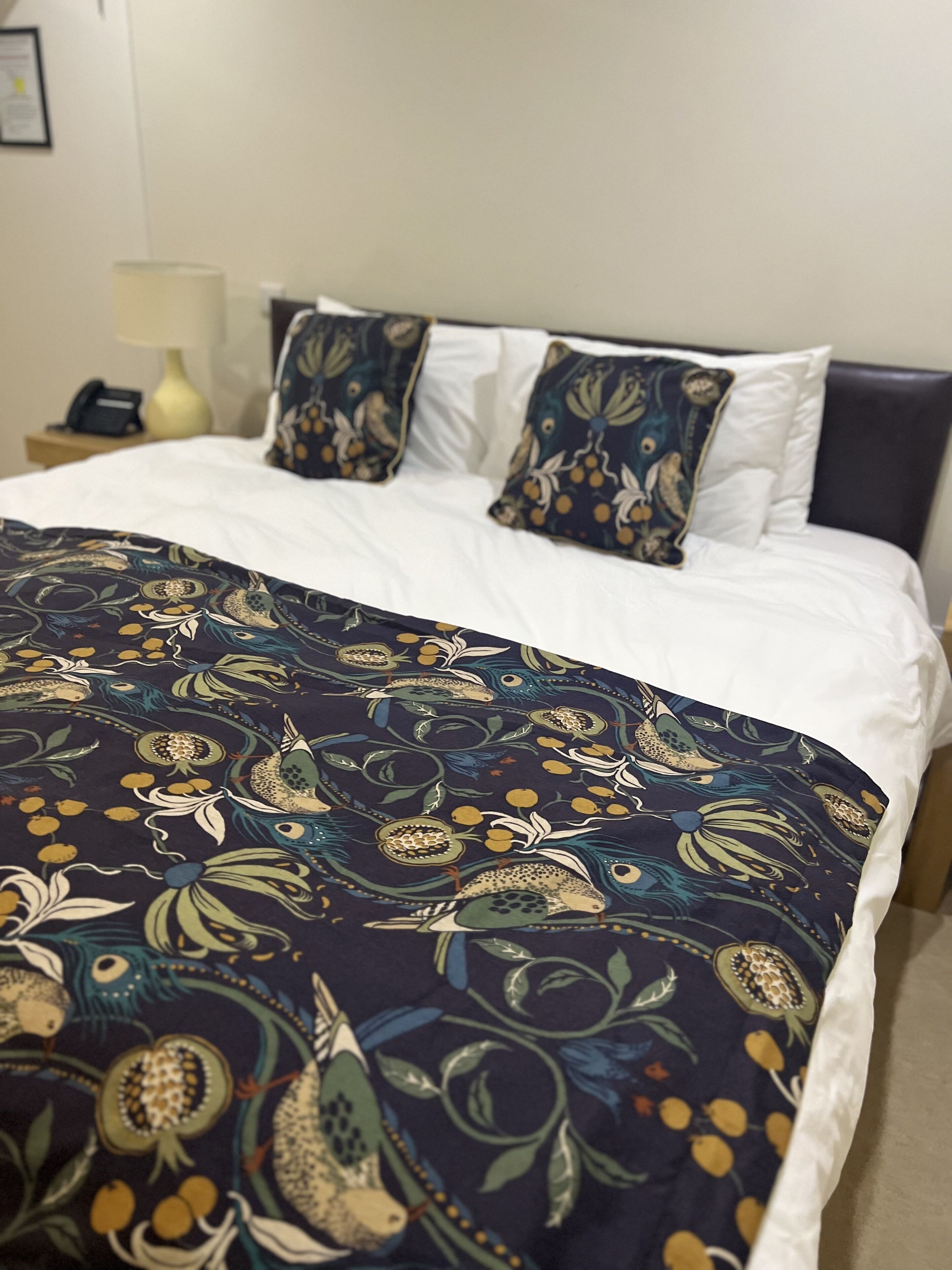 A bed with a dark blue floral bedspread and matching pillows, white sheets, and a dark headboard.