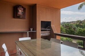 Een balkon met een glazen tafel en stoelen, een barbecue en uitzicht op de omliggende begroeiing.