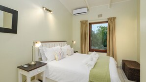 A neatly made bed with white linens, a bedside lamp, and a green throw blanket. There is a window with curtains, a small wicker stool, and a wall-mounted air conditioner. - San Lameer Villa Rentals  3014 (Southbroom)