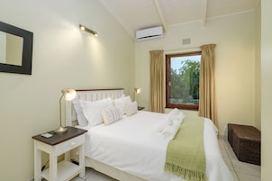 Una cama bien hecha con sábanas blancas, una lámpara de noche y una manta verde. Hay una ventana con cortinas, un taburete de mimbre pequeño y un aire acondicionado instalado en la pared.