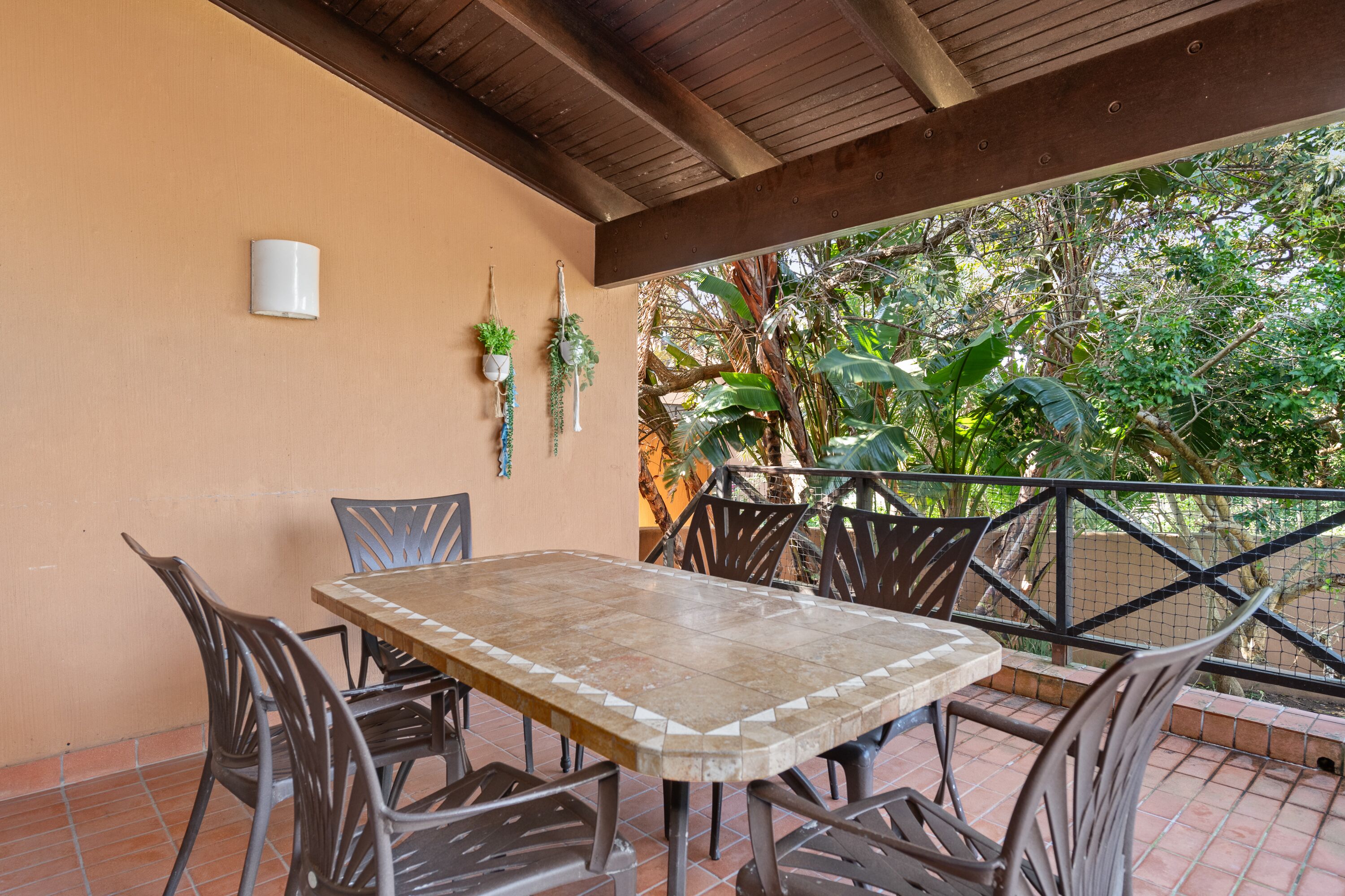 A covered patio with a tiled table and metal chairs, surrounded by a wooden ceiling and a railing.