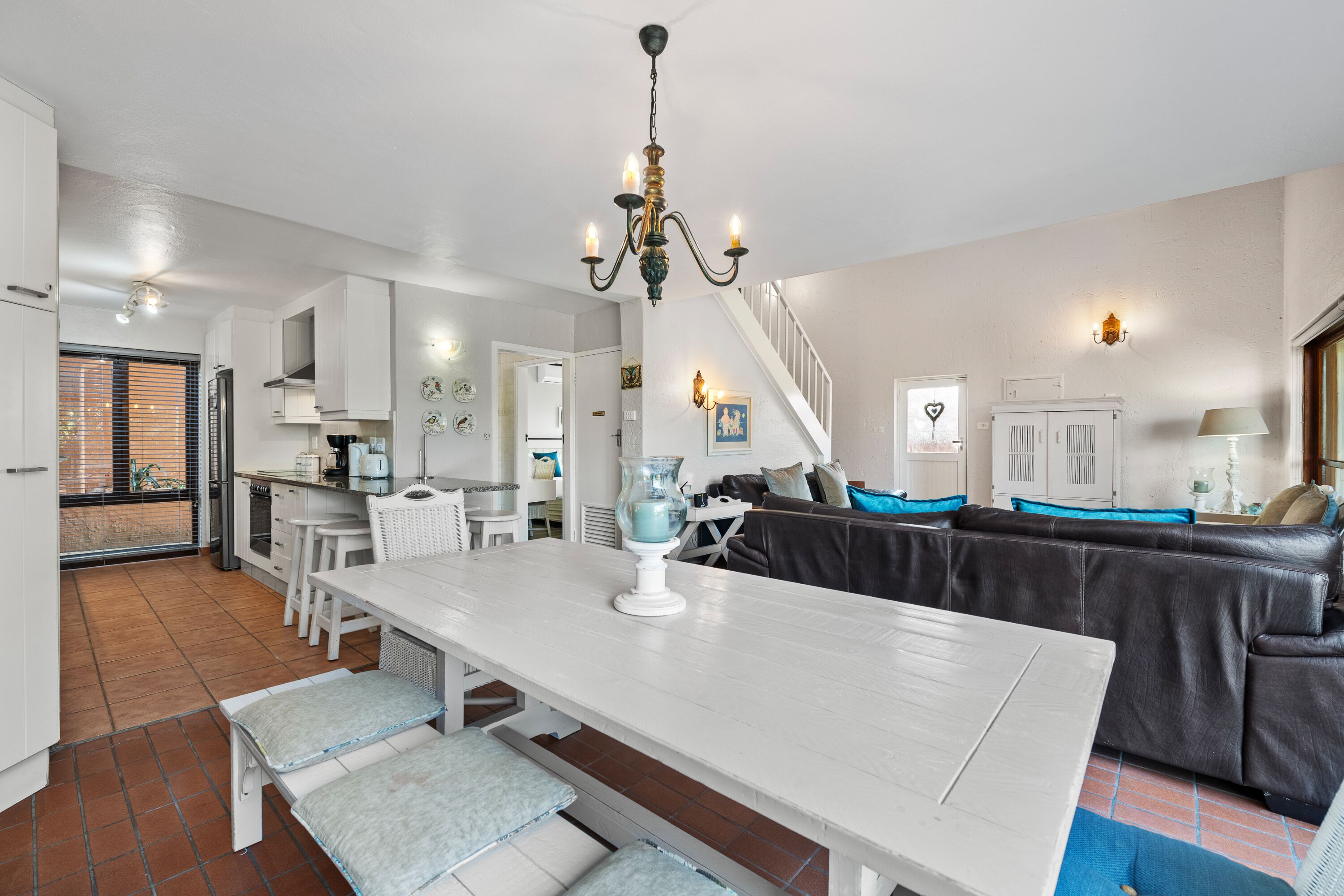 A spacious living area with a white dining table, a black leather sofa, and a staircase leading to an upper floor.