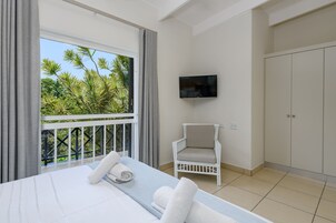 Habitación de hotel con cama, televisión, silla y balcón con vistas a palmeras.