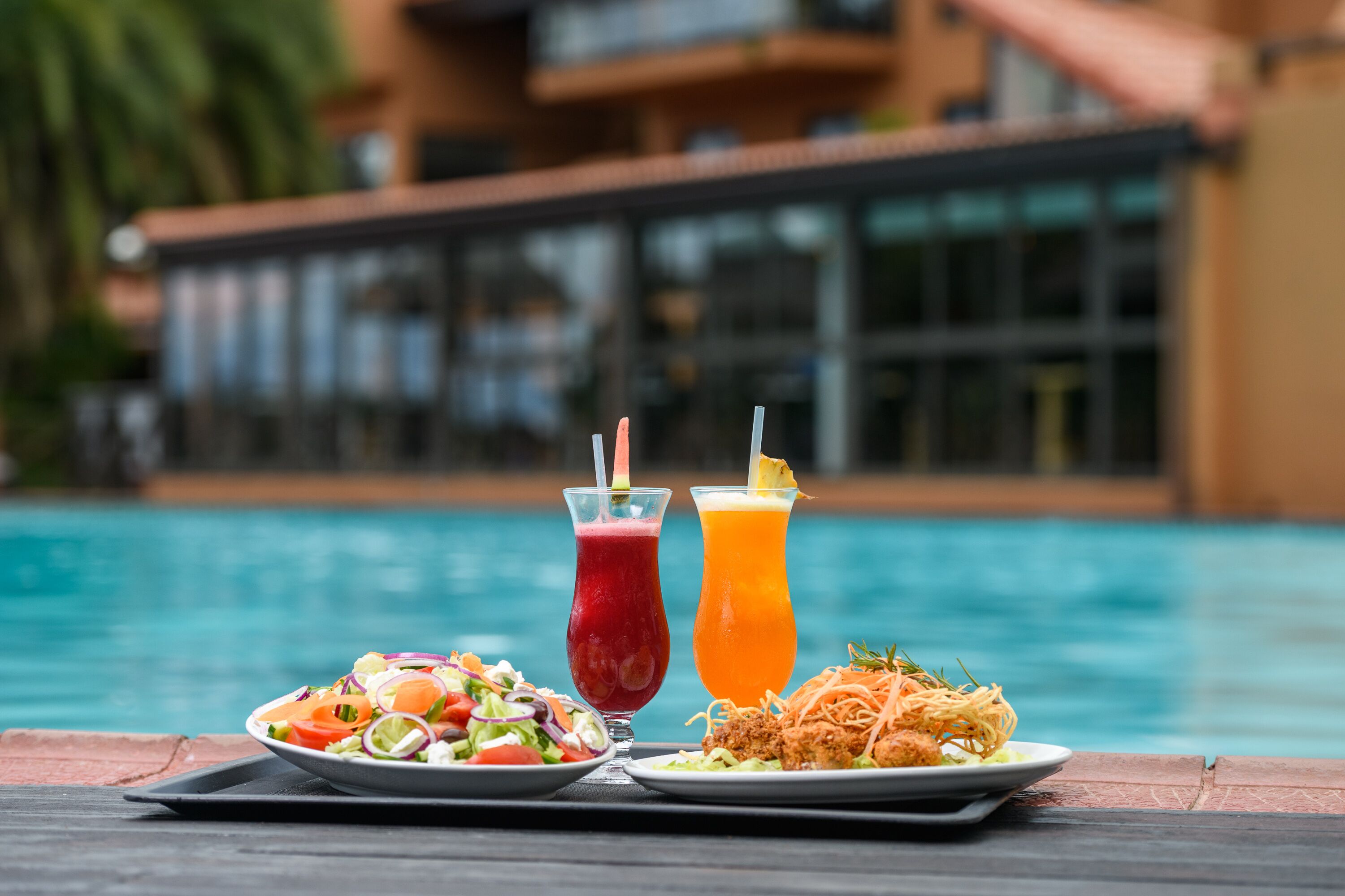 A dining table with a salad, a red drink, and a yellow drink by a poolside.