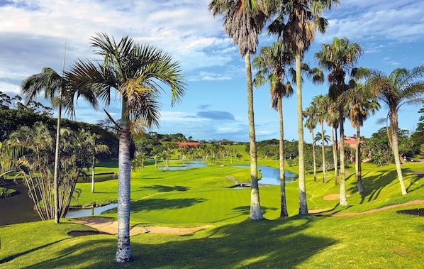 Un campo de golf con palmeras, obstáculos acuáticos y un club de campo al fondo.